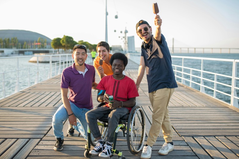 Eine Gruppe von vier Freunden steht auf einem Pier, einer von ihnen im Rollstuhl. Sie machen ein Selfie und strahlen Freundschaft und Inklusivität aus