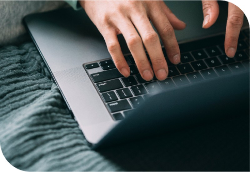 Jemand tippt auf einem Laptop, der auf einer grauen Decke liegt. Die Hände navigieren geschickt über die Tastatur, während sie die allgegenwärtige Technologie in unserem täglichen Leben repräsentieren.