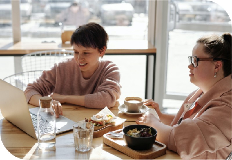 Das Bild zeigt zwei Personen an einem Tisch mit Essen, wobei eine Person mit Down-Syndrom uns zugewandt ist und die andere dem Rücken zur Kamera hat, in einem hellen, verglasten Raum.