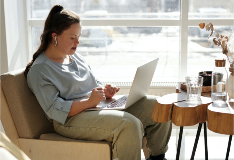 Das Bild zeigt eine Person mit Down-Syndrom, die auf einem Stuhl am Fenster sitzt, mit einem Laptop auf dem Schoß und einem runden Holztisch mit Wasser neben sich.